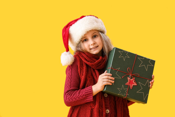Little girl in Santa hat with Christmas gift on yellow background - Fotoğraf, Görsel