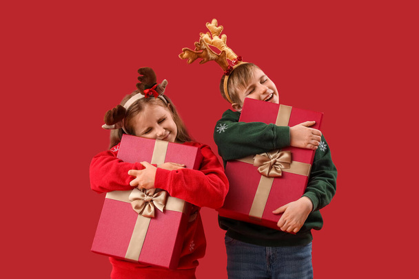 Little children in reindeer horns hugging Christmas presents on red background - Fotografie, Obrázek