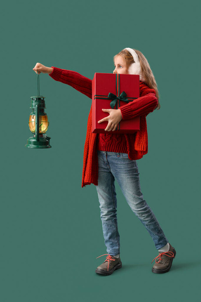 Little girl in earmuffs with Christmas gift and lantern on green background - Φωτογραφία, εικόνα
