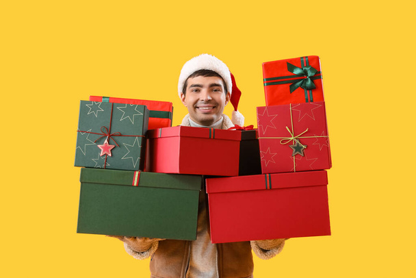Glücklicher junger Mann mit Weihnachtsmütze mit Weihnachtsgeschenken auf gelbem Hintergrund - Foto, Bild