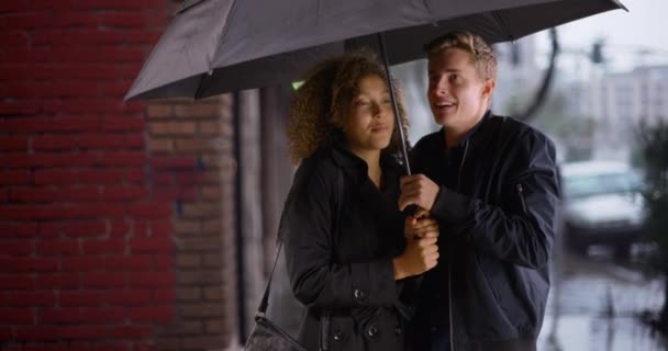 Szczęśliwy stylowy młody wieloetniczny para gospodarstwa parasol podczas deszczu - Materiał filmowy, wideo
