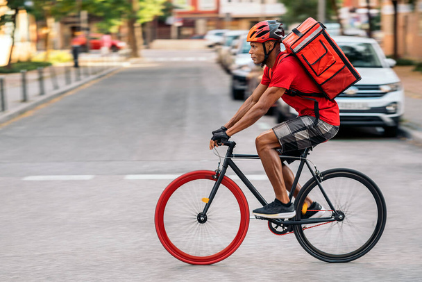 African delivery man wearing safety helmet riding his bike in the city in his way to deliver a package. - Φωτογραφία, εικόνα