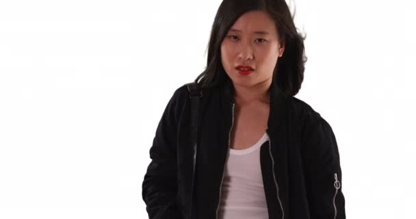 Китайка жінка в чорній куртці бомбардувальника дивиться на камеру на білому тлі. Азіатська тисячолітня, у свої 20 років, з волоссям, що віє. 4k - Кадри, відео