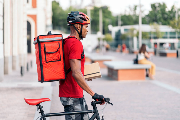 African delivery man wearing safety helmet standing with his bike in the city and holding a package. - Φωτογραφία, εικόνα
