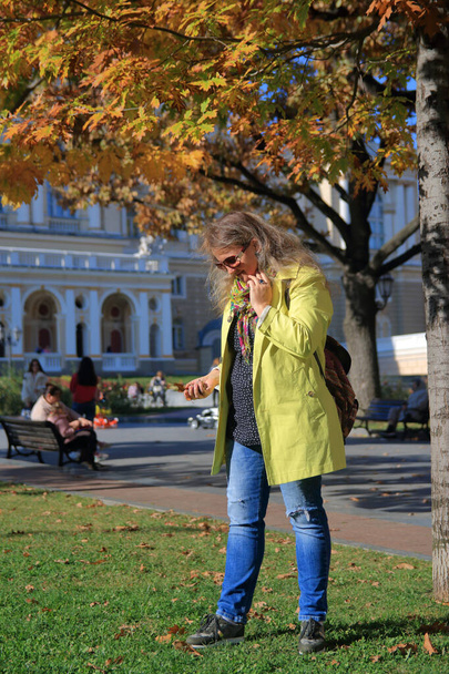 A foto foi tirada na cidade de Odessa, na Ucrânia. A imagem mostra uma jovem mulher em uma capa de chuva amarela à procura de bolotas na grama verde de um parque de outono. - Foto, Imagem