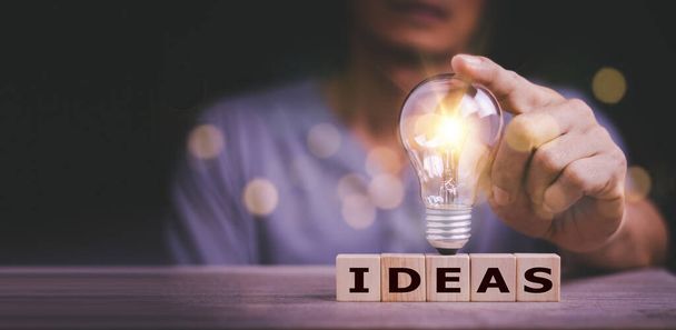Hand anfassen Glühbirne auf Holzblock mit Word Ideas, neues Ideenkonzept mit Innovation und Inspiration, innovative Technologie in Wissenschaft und Kommunikationskonzept. - Foto, Bild