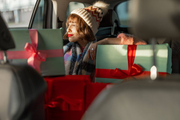 Mladá žena oblečená na svátky jezdí autem na zadním sedadle s krásně zabalenými vánočními dárky. V očekávání zimních prázdnin, přípravy a nakupování - Fotografie, Obrázek