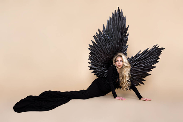 Stunning blonde woman in elegant long black dress and big wings is posing in studio. Luxury evening fashion. Glamour fashion model. Woman with wings. Black Angel. - Zdjęcie, obraz