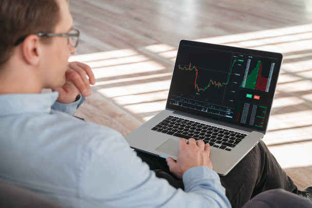 Close up shot of trade investor sitting with laptop on knees, analyzing candlestick diagram with stock market app, trying to predict actions of money flow, waiting for best moment to buy crypto - Foto, Imagen