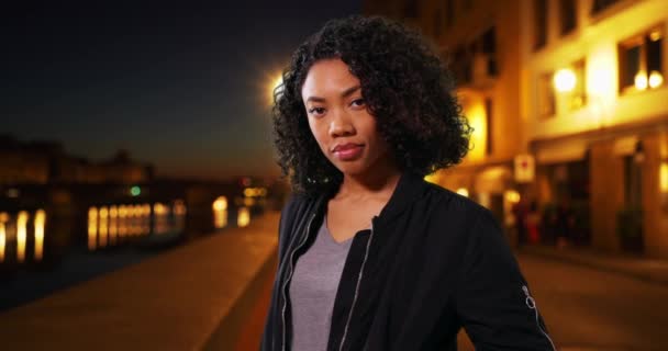 Beautiful African American Millennial woman standing outdoors in Florence on cool evening. Female tourist wearing black bomber jacket looking at camera at night. 4k - Footage, Video