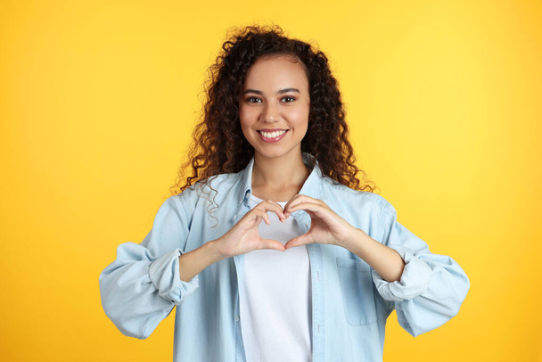Happy young African-American woman making heart with hands on yellow background - Photo, image