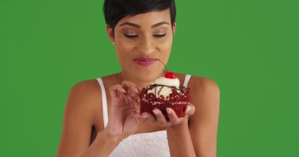 Закройте привлекательную черную женщину, которая ест большой красный бархатный кекс на зеленом экране. На зеленом экране клавиатура или композиция. - Кадры, видео