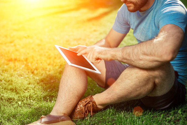 Hipster młody chłopak siedzi w parku i przy użyciu nowoczesnego tabletu cyfrowy, mężczyzna blogger za pomocą tabletu pc poza social networking koncepcja. Rozbłysk słoneczny - Zdjęcie, obraz