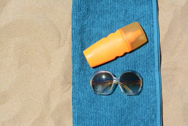 Soft blue beach towel with bottle of sunblock and sunglasses on sand, flat lay. Space for text - Foto, Bild