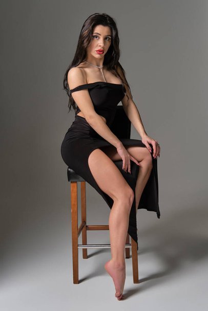 Fotografie mladé atraktivní brunetky běloška sexy žena sedí na židli na sobě erotické koktejlové černé šaty. Šedé pozadí, studio pořízené vysoce kvalitní obraz - Fotografie, Obrázek