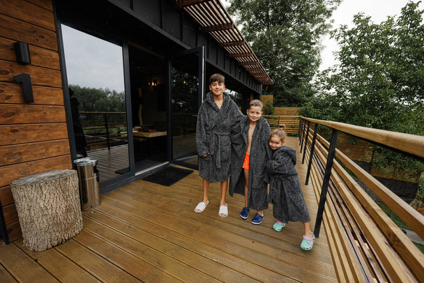 Three kids in bath robe stand in wooden terrace of modern house. - Foto, afbeelding
