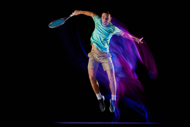 Movements in neon glow. Active male tennis player in sportwear playing tennis isolated over dark background in mixed purple neon light. Concept of motion, speed, professional sport. - Fotoğraf, Görsel