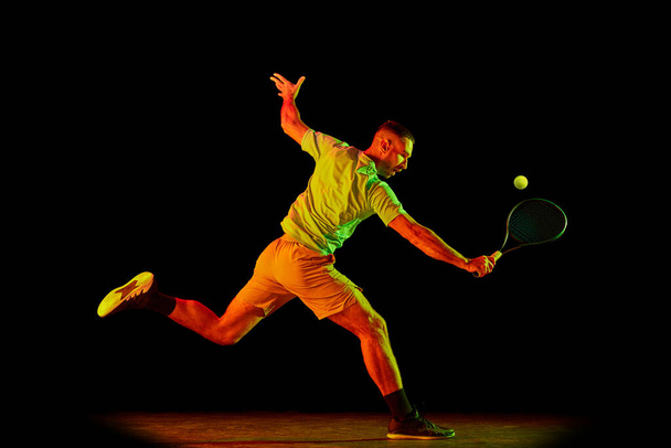 Athlete playing tennis. Young sportive man, professional tennis player in motion, action isolated over dark background in neon light. Concept of professional sport, competition, skills. - Valokuva, kuva