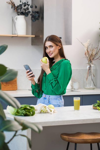 An attractive brunette woman holds a green apple and smiles at the camera while standing at the kitchen table. Healthy lifestyle, vegetarianism, - Foto, Imagen