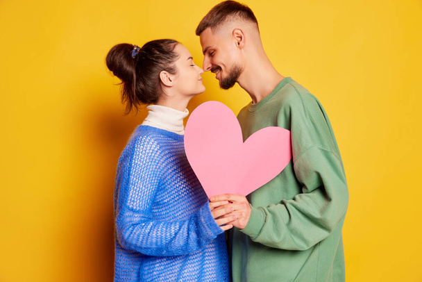 Portrait of beautiful young couple, man and woman posing nose to nose with paper heart isolated over yellow background. Concept of love, relationship, Valentines Day, emotions, lifestyle, dating - Foto, imagen