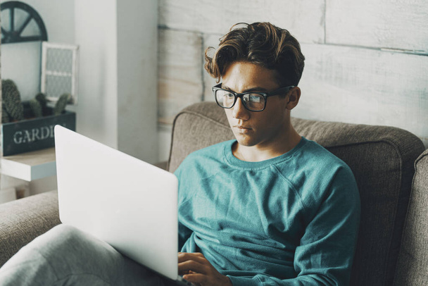 Egy diák fiú otthon laptopot használ, és koncentrált arckifejezéssel ül a kanapén. Fiatal tinédzser férfi szemüvegben, aki számítógépen dolgozik. Online oktatási cél és szabadidős technológia - Fotó, kép