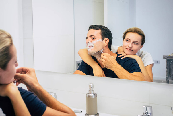 Fekete-fehér fiatal pár szórakozik otthon a fürdőszobában. Férfi borotválkozás szakáll a reggeli rutin és a lány mosolyog. Az együttélés fogalma egy kapcsolatban. Boldog emberek beltéri szabadidő - Fotó, kép