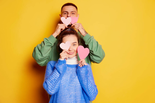 Portrait of beautiful young couple, man and woman posing with paper hearts isolated over yellow background. Funny faces . Concept of love, relationship, Valentines Day, emotions, lifestyle, date - Zdjęcie, obraz
