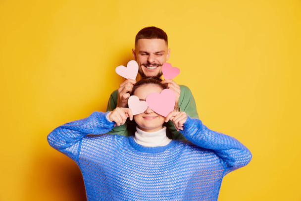 Portrait of beautiful young couple, man and woman posing with paper hearts isolated over yellow background. Cheerfully smiling. Concept of love, relationship, Valentines Day, emotions, lifestyle - Photo, Image