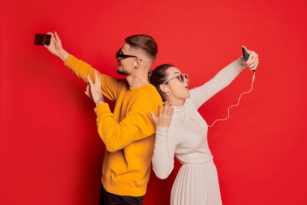 Portrait of happy young couple, man and woman, standing back to back and having video call on phone isolated over red background. Concept of love, relationship, Valentines Day, emotions, lifestyle - Photo, image