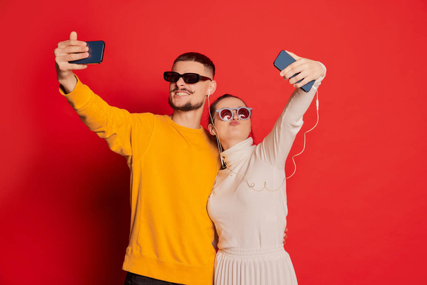 Portrait of happy young couple, man and woman, posing, taking selfie with phone isolated over red background. Looking happy. Concept of love, relationship, Valentines Day, emotions, lifestyle - Foto, afbeelding