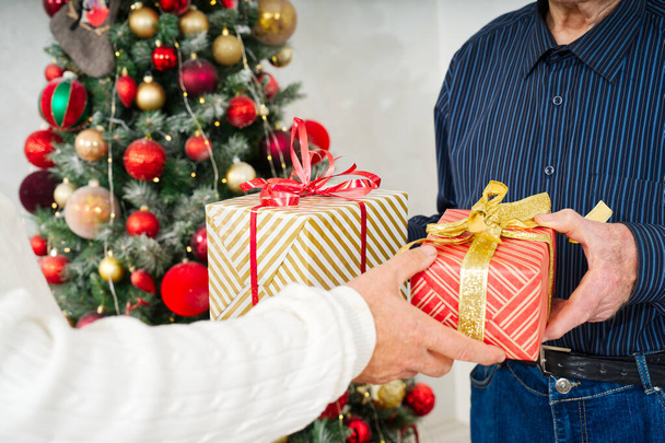 Ajándékcsere a díszített karácsonyfa előtt. Felnőtt kezek közelsége, akik karácsonyi ajándékot tartanak csomagolópapírban. Romantikus nap. Téli ünnepek. - Fotó, kép