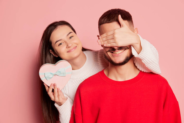 Portrait of young couple, beautiful woman covering mans eyes and giving present isolated over pink background. Holiday surprise. Concept of love, relationship, Valentines Day, emotions, lifestyle - Photo, image