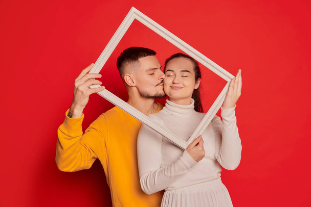 Portrait of happy young couple, man and woman, posing with picture frame isolated over red background. Giving a kiss to a girl. Concept of love, relationship, Valentines Day, emotions, lifestyle - Foto, afbeelding