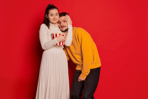 Portrait of happy young couple, man and woman, posing isolated over red background. Watching horror movie together, hugging . Concept of love, relationship, Valentines Day, emotions, lifestyle - Фото, изображение