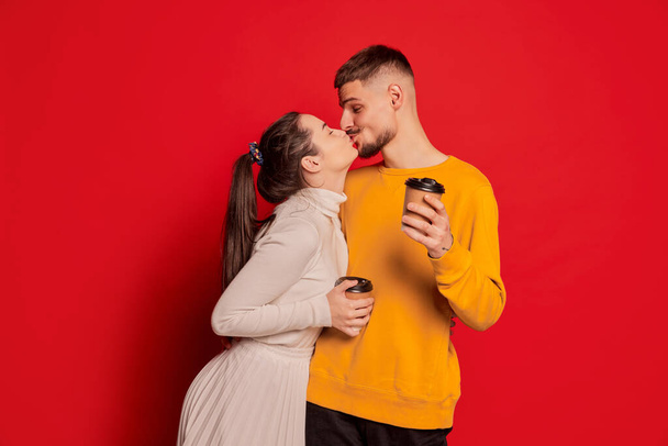Portrait of happy young couple, man and woman, posing, kissing isolated over red background. Coffee break. Concept of love, relationship, Valentines Day, emotions, lifestyle, leisure time - Photo, image