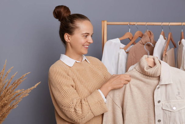 Attractive smiling woman with dark hair and bun hairstyle holding clothes on racks in modern boutique, choosing attire, trying to make stylish trendy look, expressing happiness. - Foto, imagen