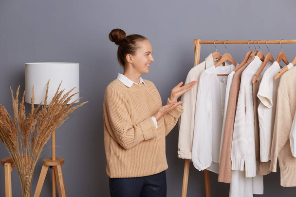 Portrait of happy attractive woman stylist wearing beige knitted sweater standing and showing shelves with clothes, female designer presenting her new collection, expressing positive emotions. - Foto, Imagem