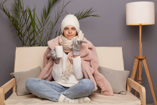 Portrait of winsome sad upset cold woman sitting on the sofa at home drinking hot tea and using smart phone, female wearing winter coat, hat and mittens. - Fotoğraf, Görsel