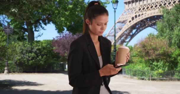 Young professional woman on business trip in Paris sending text message under Eiffel Tower while drinking coffee. Millennial businesswoman using cell phone while meeting colleague in France. 4k - Footage, Video