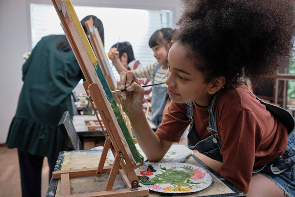 African American κορίτσι επικεντρώνεται στην ακρυλική ζωγραφική έγχρωμη εικόνα σε καμβά με τους μαθητές ομάδα στην αίθουσα τέχνης, δημιουργική μάθηση με ταλέντα και δεξιότητες στην εκπαίδευση δημοτικό σχολείο στούντιο. - Φωτογραφία, εικόνα