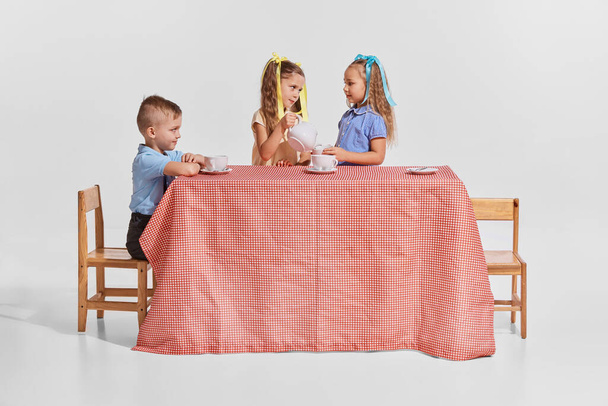 Πορτρέτο τριών παιδιών, αγοριών και κοριτσιών που κάθονται στο τραπέζι πάνω από γκρι φόντο. Ώρα για τσάι. Επικοινωνία. Έννοια της παιδικής ηλικίας, δημιουργικότητα, ρετρό μόδα, φιλία, τέχνη - Φωτογραφία, εικόνα