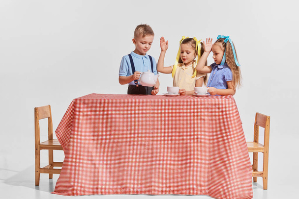 Portrait of three children, boy pouring tea into girls cup isolated over gray background. Breakfast. Spending time together. Concept of childhood, creativity, retro vintage fashion, friendship, art - Photo, image