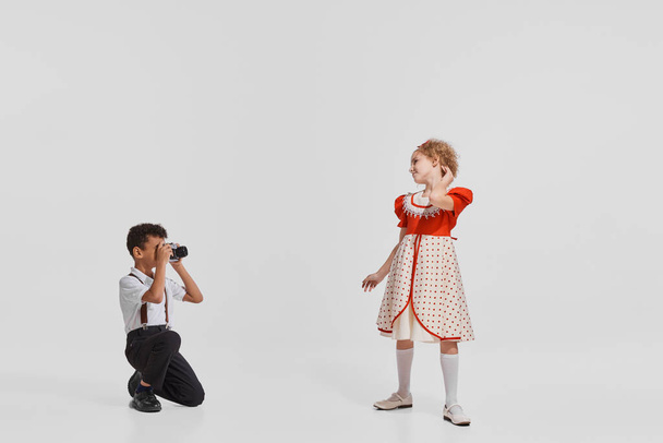 Portrait of little children, boy taking photo of pretty girl in cute old-fashioned dress isolated over grey background. Concept of childhood, creativity, retro vintage fashion, friendship, art - Zdjęcie, obraz