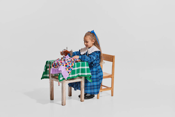 Portrait of pretty little girl in cute blue dress sitting at the table and sewing isolated over grey background. Concept of childhood, creativity, retro style, vintage fashion, friendship, art - Φωτογραφία, εικόνα