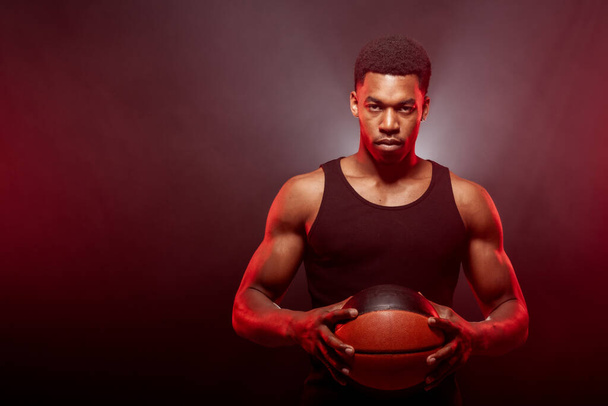 Basketball player side lit with red color holding a ball against hazy background. Serious concentrated african american man. - Фото, зображення