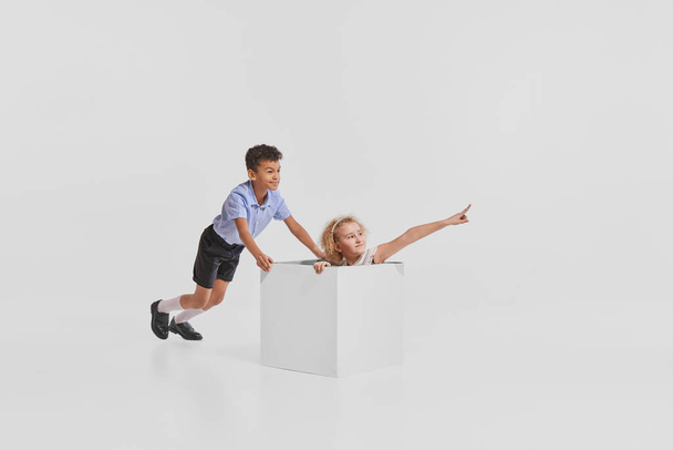 Portrait of two children, girl playing together, girl sitting in box isolated over grey studio background. Sailing. Concept of childhood, creativity, retro style, vintage fashion, friendship, art - Photo, Image