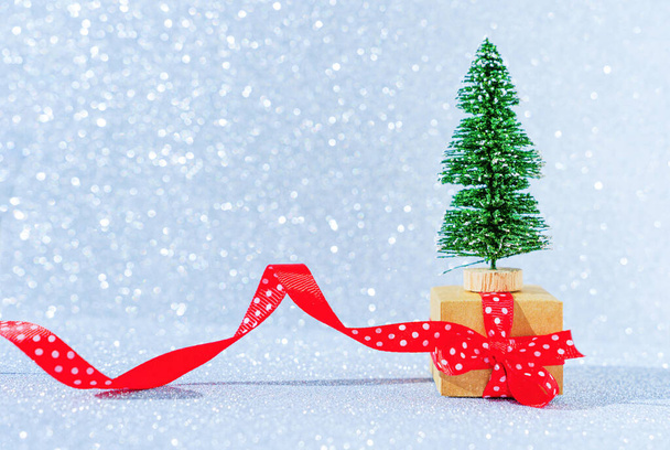 Miniature gift box wrapped with red ribbon and placed under a toy christmas tree against a sparkling background. - Foto, imagen