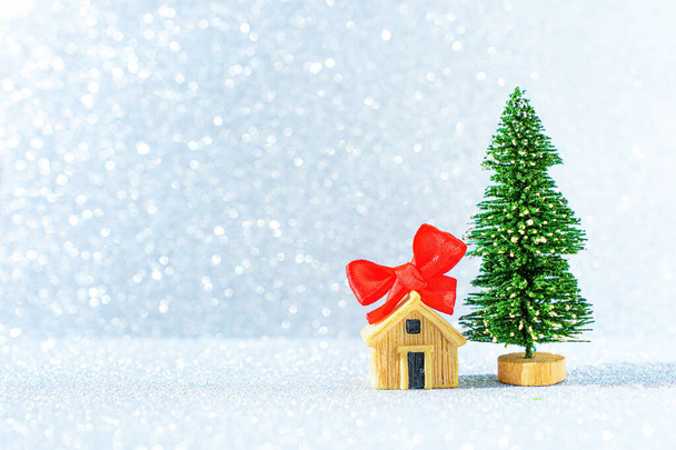 Toy new year tree and miniature house figurine decorated with a red bow against a glittering winter background. - Fotó, kép