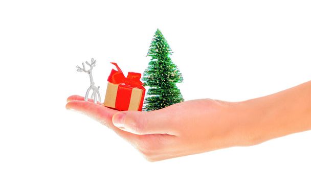 Hand holding a toy christmas tree, miniature deer figurine small gift box with red bow isolated on white background. Offering best winter holidays props and presents. - Photo, image