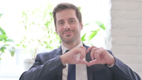 Αγαπώντας νεαρός ενήλικος επιχειρηματίας δείχνει σχήμα καρδιάς από τα χέρια στο γραφείο - Πλάνα, βίντεο